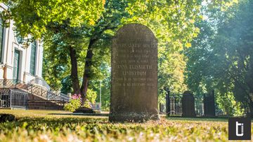 An old gravestone under trees with warm sunlight through green leaves.