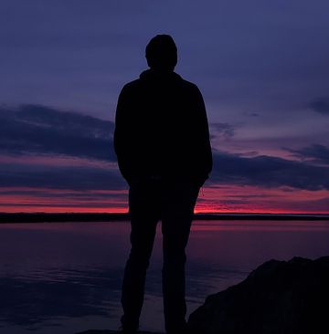 A silhouette of a man looking out onto a vibrant purple and pink sunset above a peaceful sea.